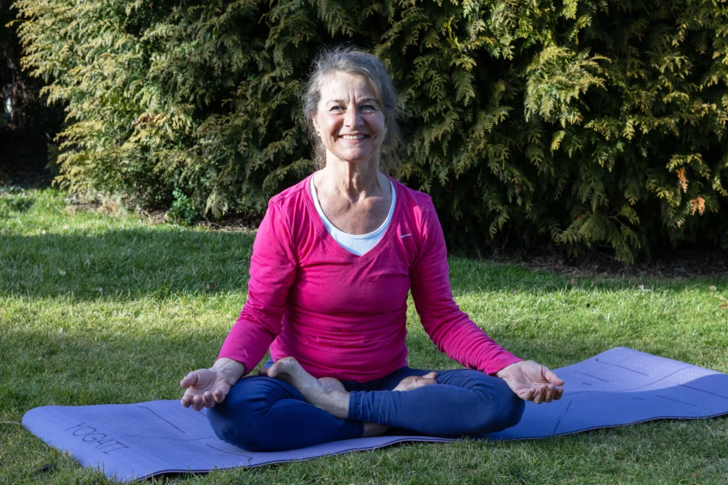Das Bild zeigt eine Frau sitzend in Yoga Pose in einer Wiese